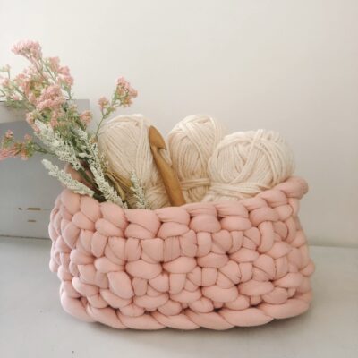 A crocheted basked is sitting on a white table. The basket made with very thick, pink yarn. Inside the basket sits 3 skeins of white yarn, a large crochet hook, and pink flowers.