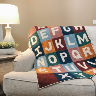 A multi-colored alphabet blanket is draped over a white couch. There is a white lumped turned on in the corner. The photo looks cozy.