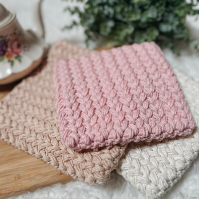 A stack of three crochet trivets sit in a pile. A white trivet is on the bottom, a tan trivet is in the middle, slightly to the left, and a pink trivet sits on top.